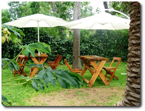La foto muestra mesas y sillas bajo sombrillas sobre el césped y entre los árboles para disfrutar de desayunos, mateadas y charlas en contacto con la naturaleza.